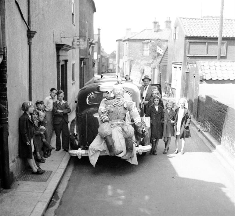 Photograph. Children and figure attached to car on Vicarage Street. (North Walsham Archive).