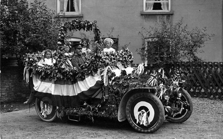 Photograph. Decorated car ready for the Coronation Procession, 1911, Palmer's Yard, Vicarage St., North Walsham. Driver has Royal Norfolk Regt. cap badge. (North Walsham Archive).